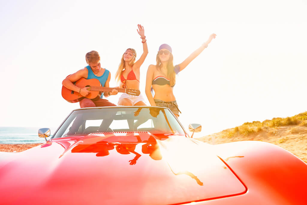Three teenagers singing and dancing while sitting in a convertible car.