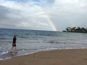 Jen Cook walking along Hawaiian Beach with Rainbow in background.