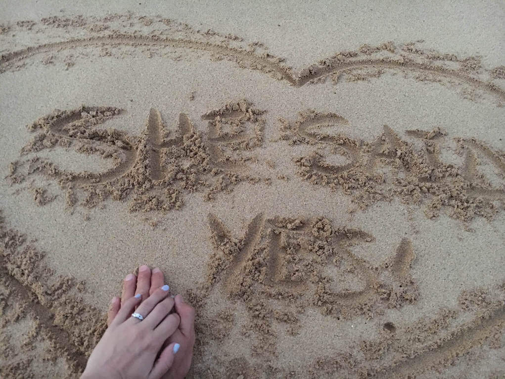 Heart drawn in sand with "she said yes" in the middle. A male hand and female hand are intertwined with an engagement ring featured.